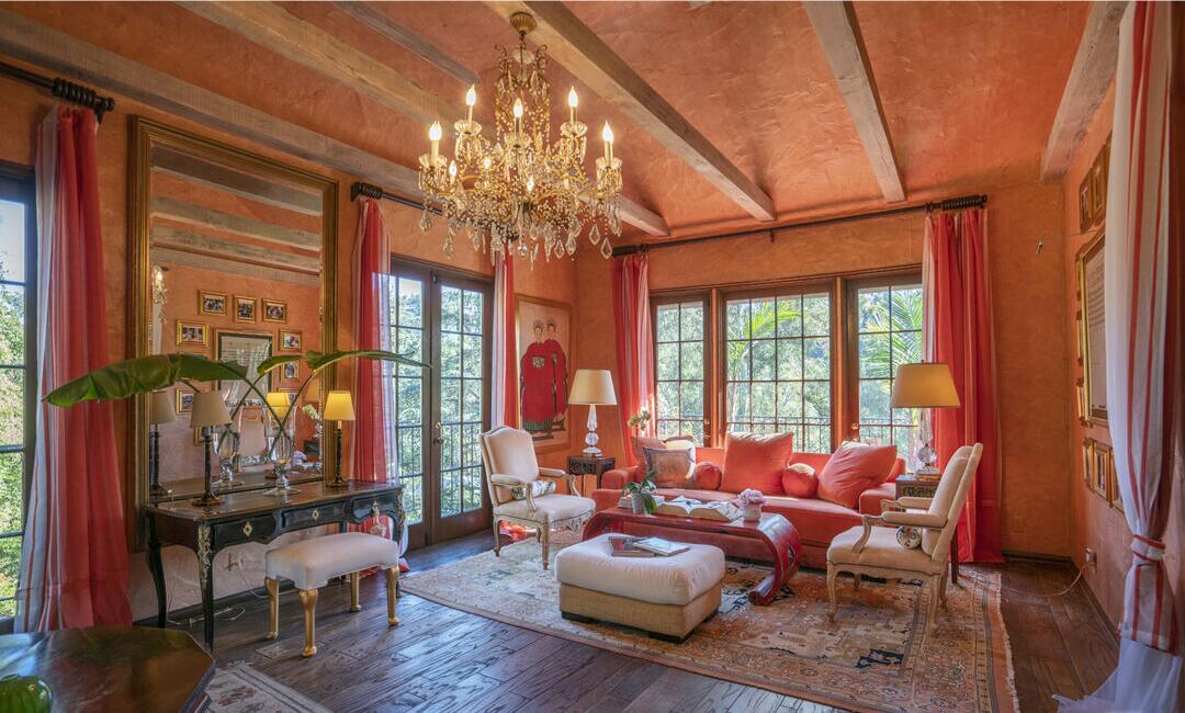 A living room in orange shades with a chandelier, furniture and windows overlooking foliage.