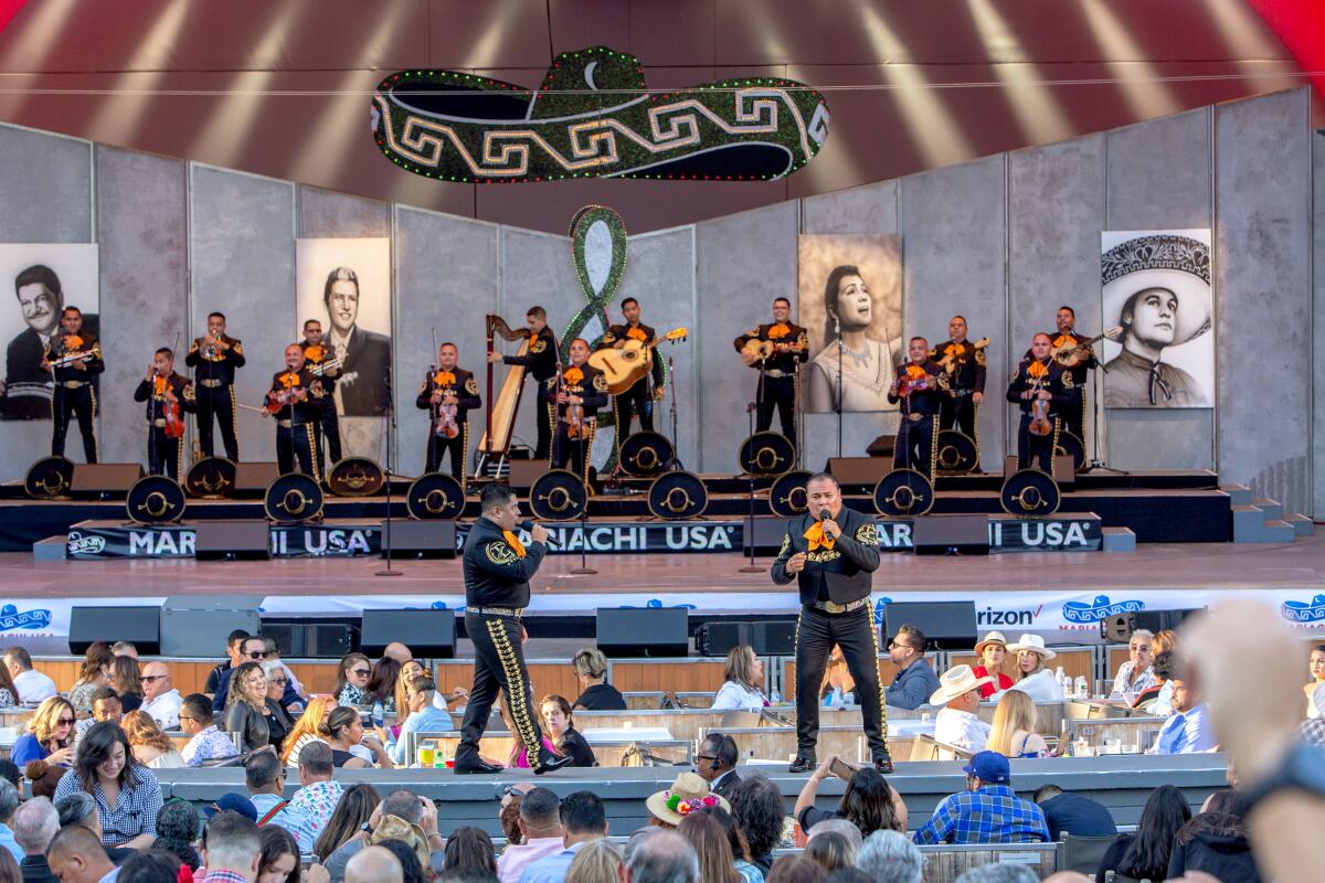 Two mariachi singers perform on stage close to audience while a mariachi band plays on a podium in the background.