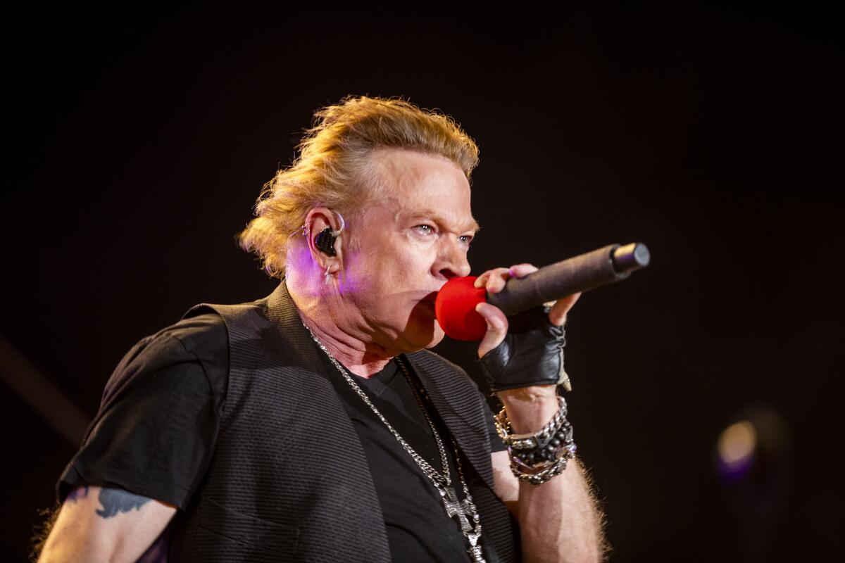 Axl Rose wears a black shirt and vest as he sings into a red-capped microphone while onstage.