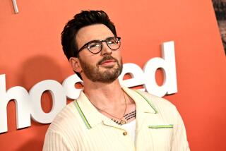 Chris Evans poses in glasses and a cream collared shirt against an orange background.