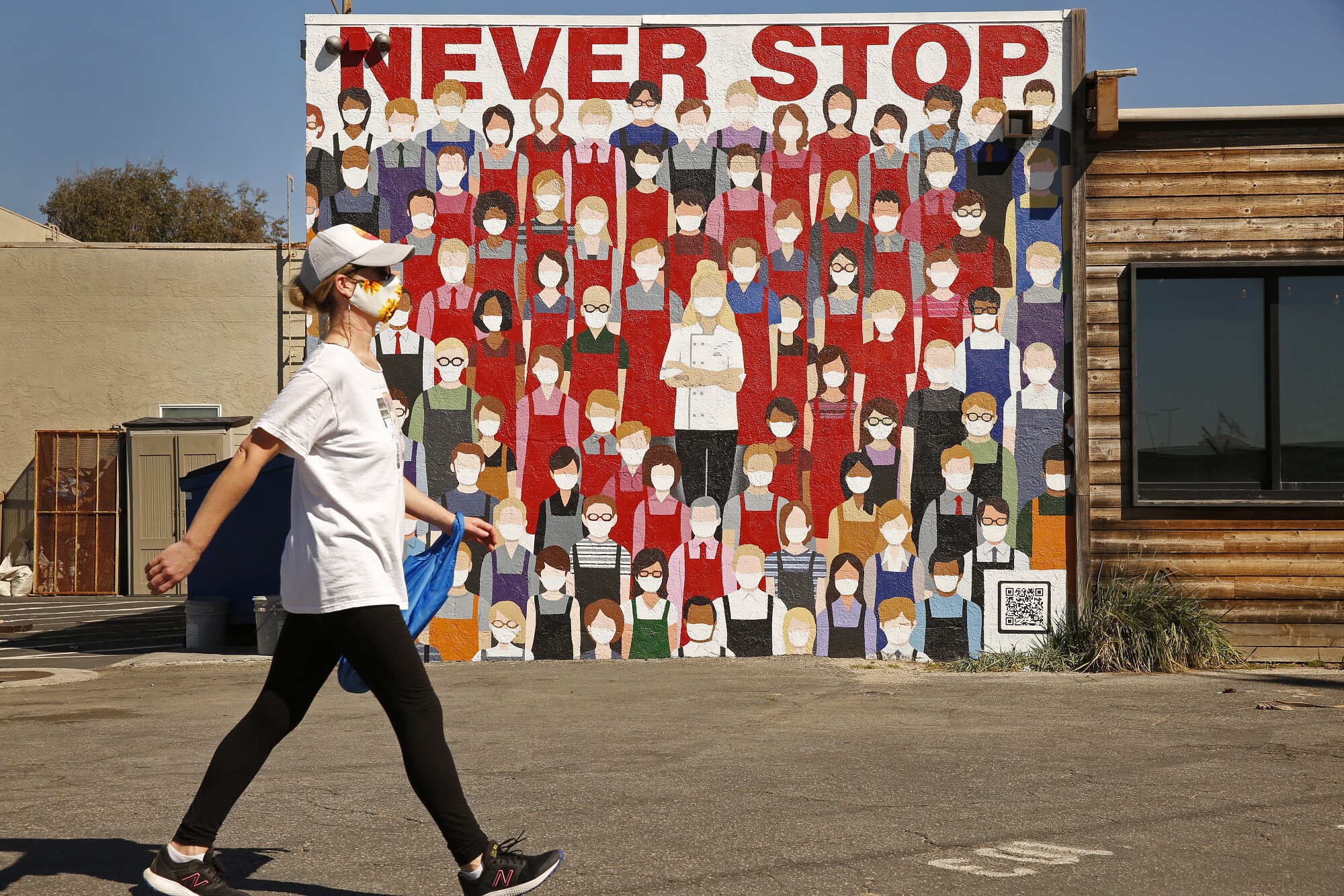 A woman walks past a mural of a crowd of people in masks