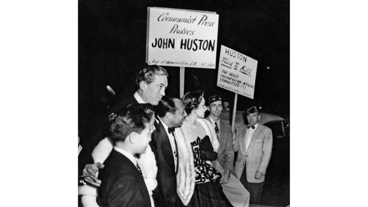 John Huston stands with his entourage in formal attire as protesters accuse him of being a communist sympathizer.