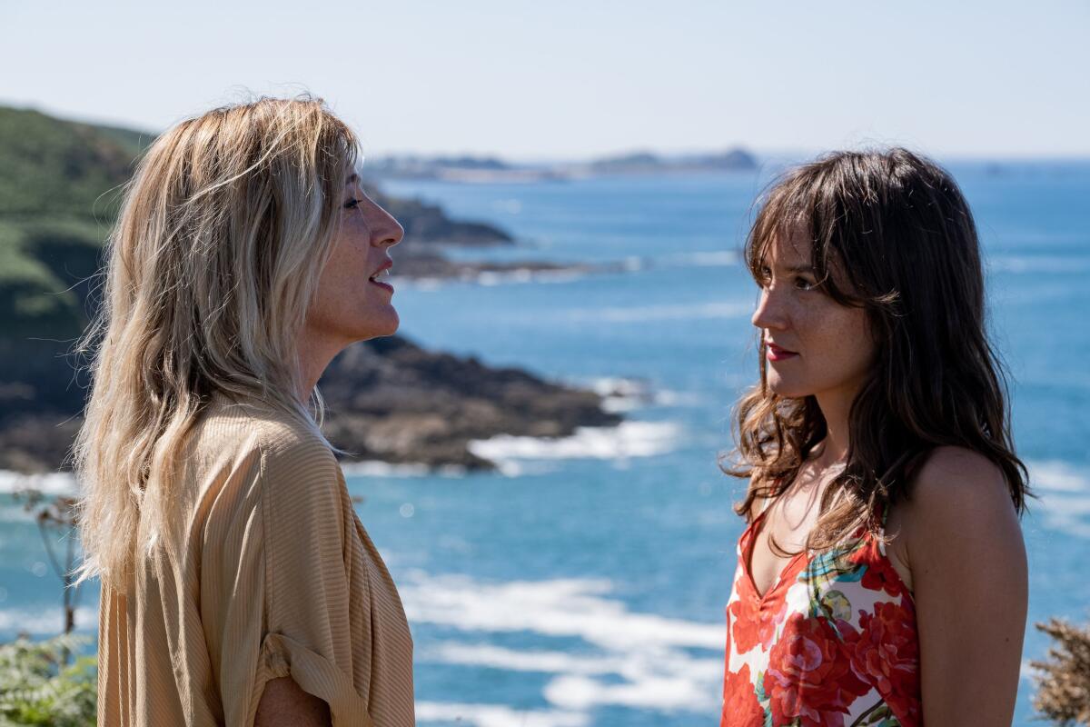 Two women face each other and talk. In the background is an ocean coastline.