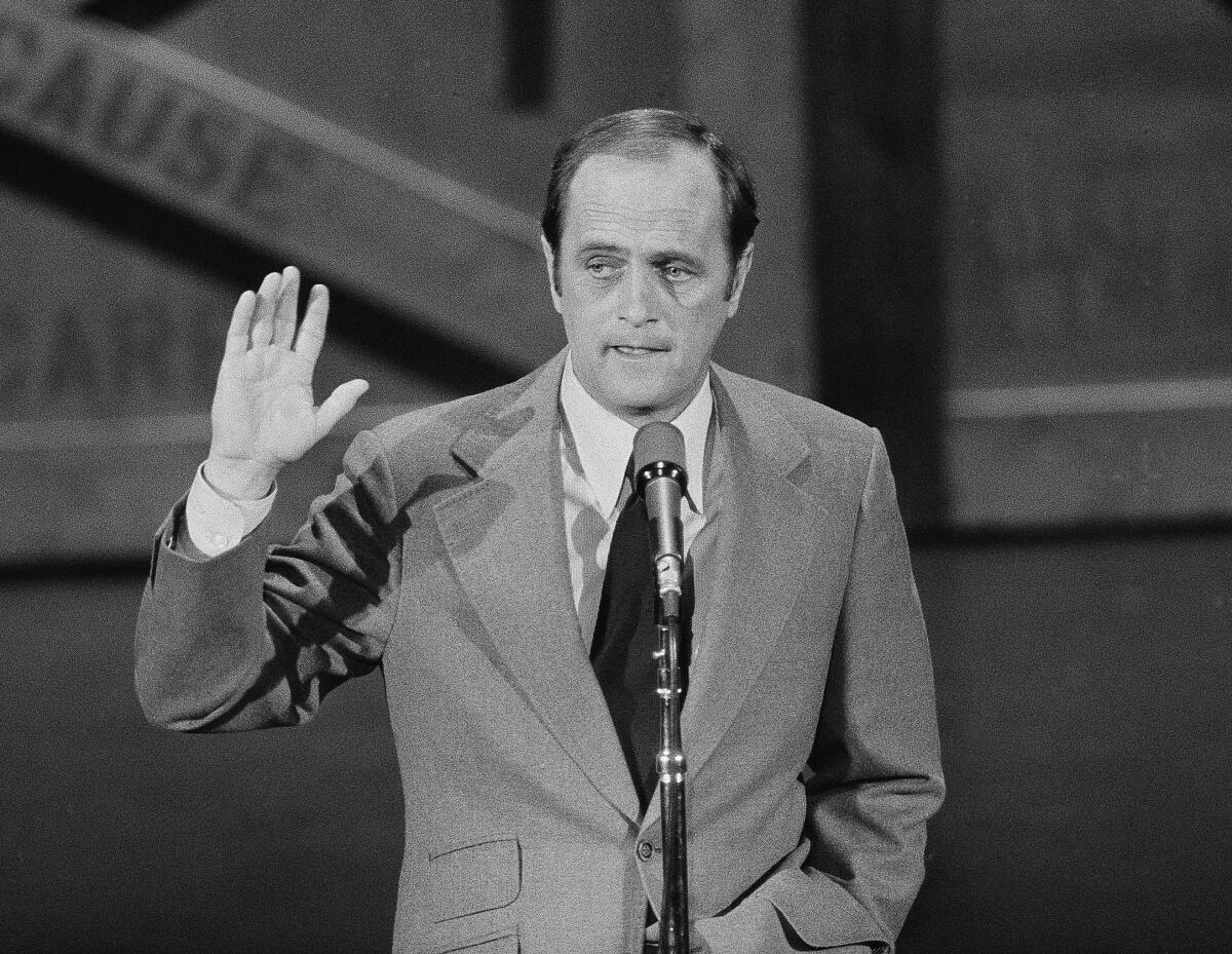 Bob Newhart standing onstage at a microphone, holding up one hand