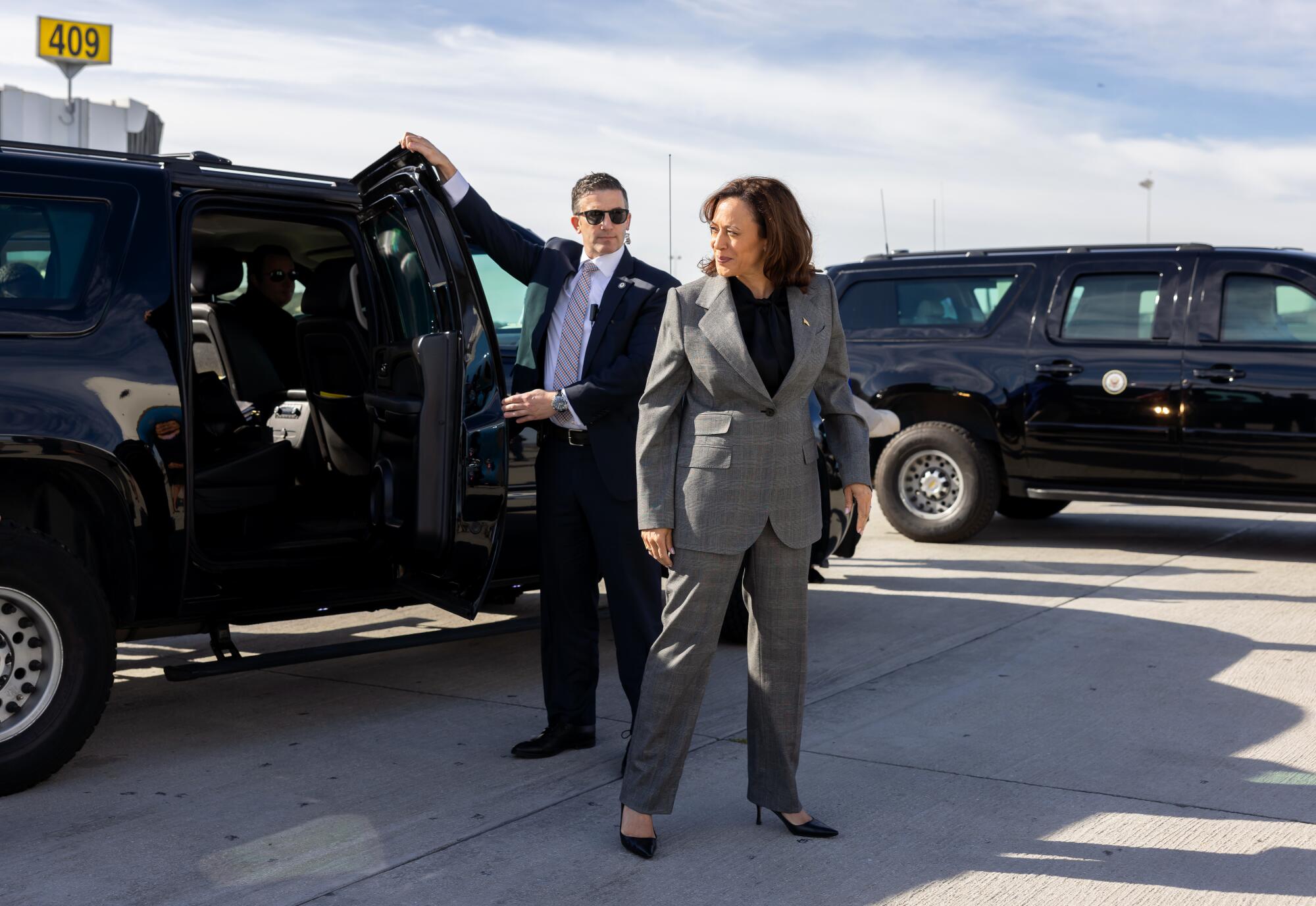 Vice President Kamala Harris stands outside a black SUV with a door opened by a man. Another black SUV is in the background.