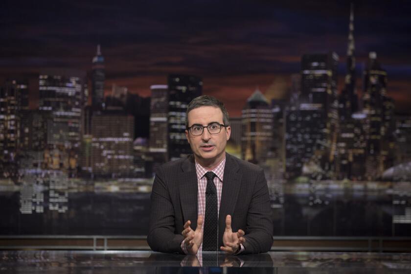 John Oliver sitting behind a desk with the New York skyline behind him