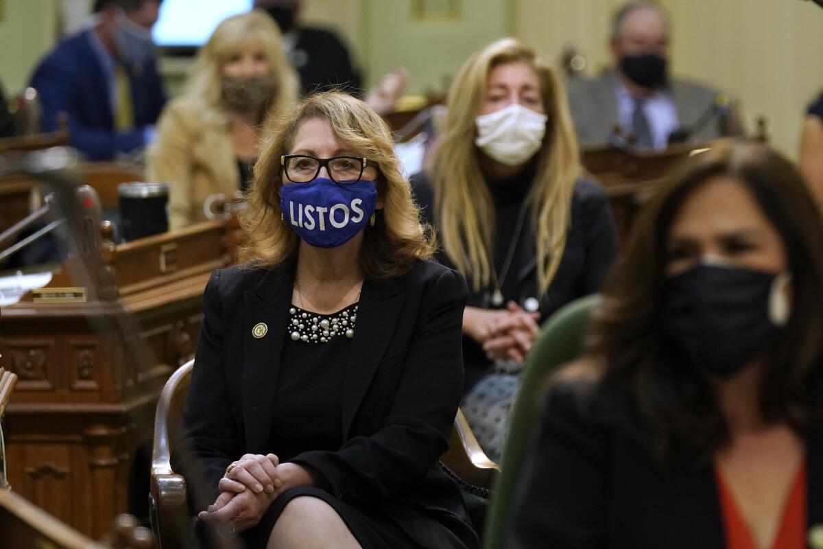 A legislator sits with her hands clasped in the Assembly chambers, wearing a blue mask that says Listos