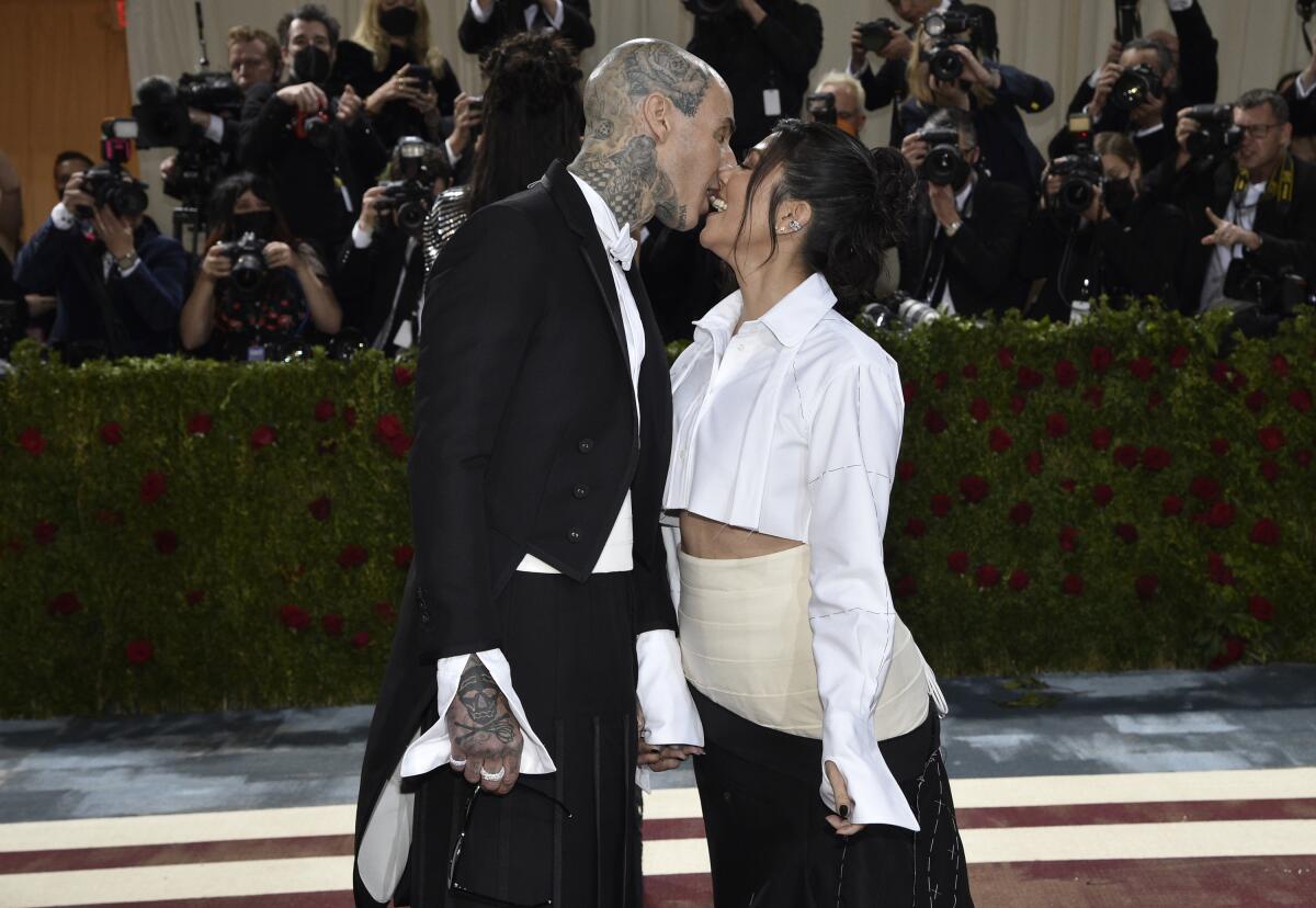 Travis Barker kisses Kourtney Kardashian in front of a bank of photographers who are standing behind a low hedge
