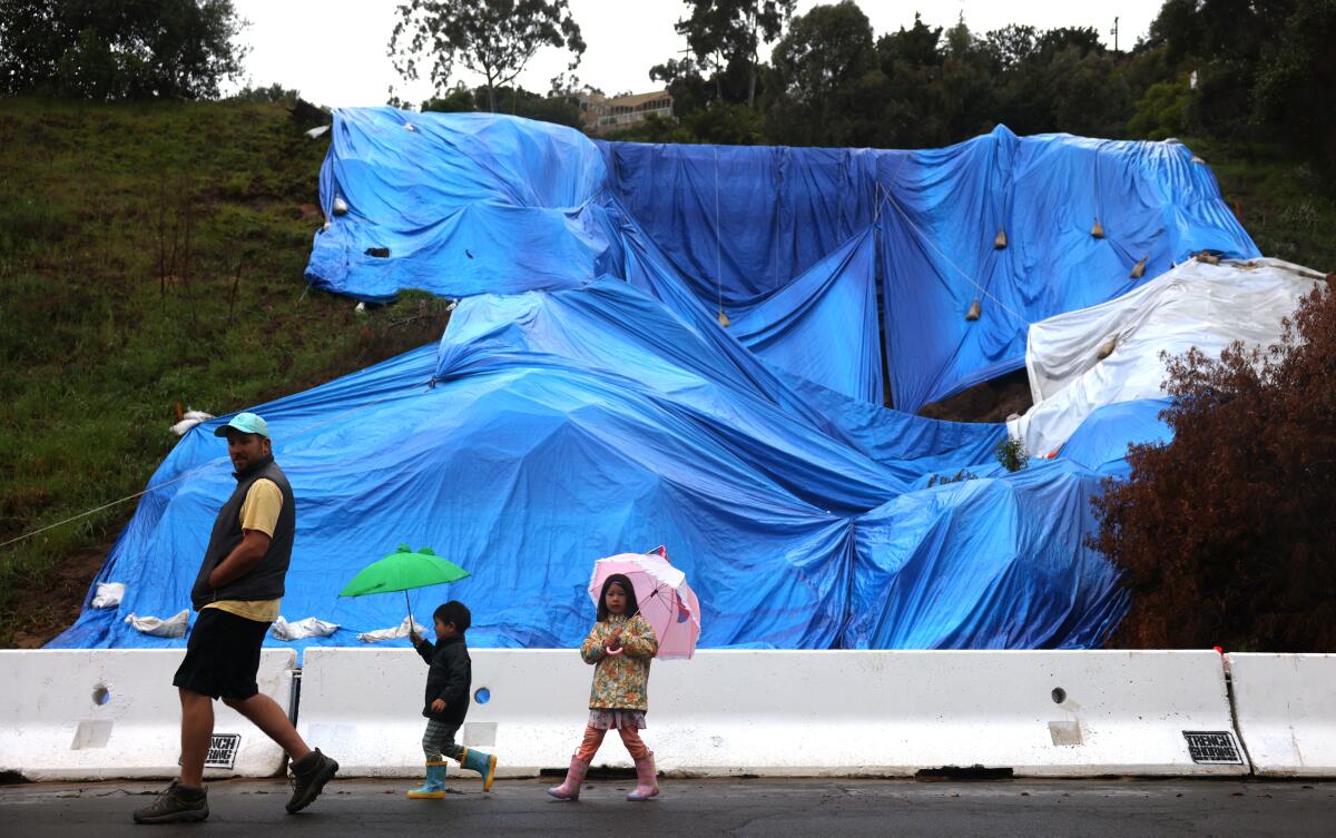 A man and two small children with umbrellas walk single-file past a damaged hillside covered in blue and white tarps