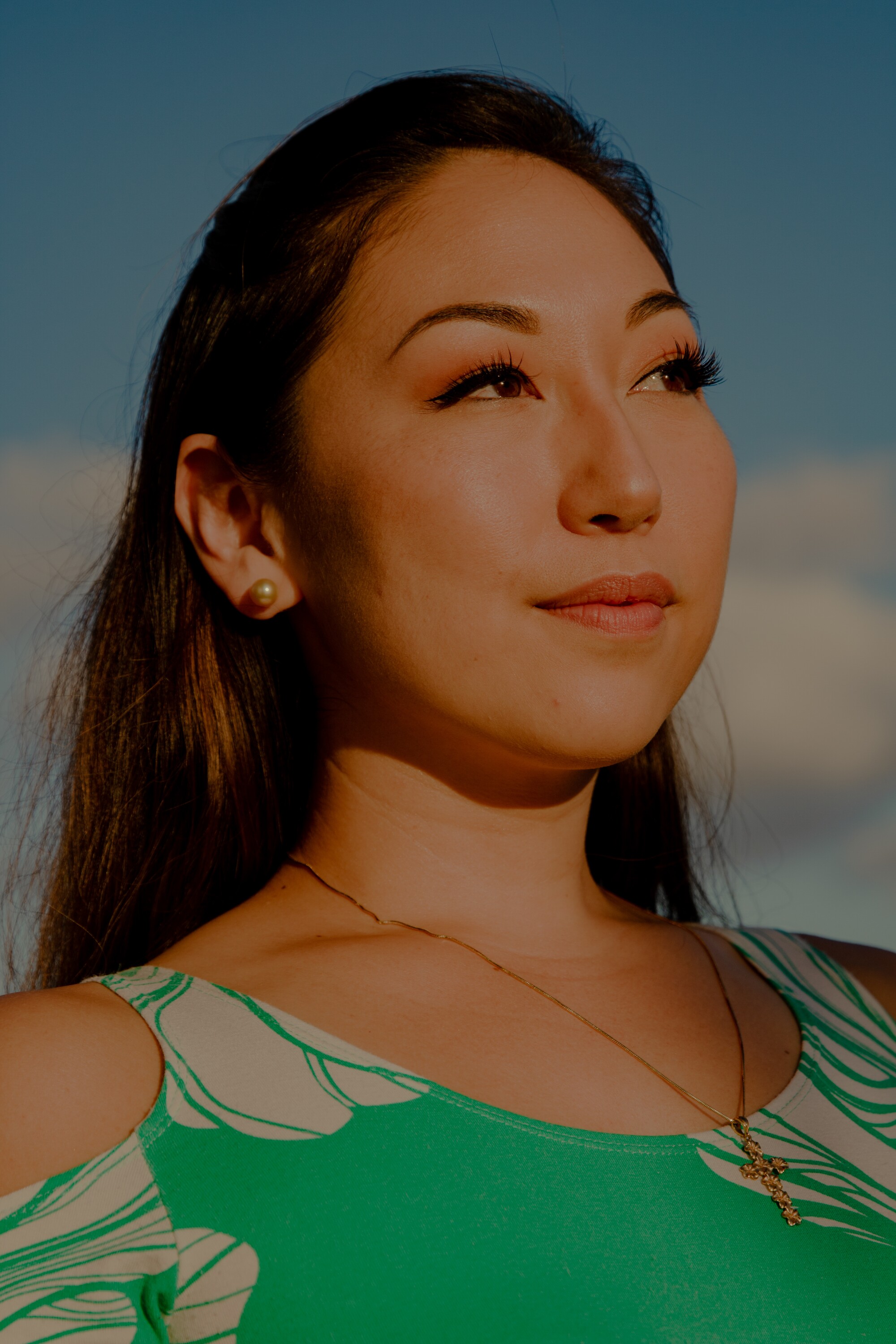 A woman wearing a green patterned top and a cross around her neck while looking off into the distance