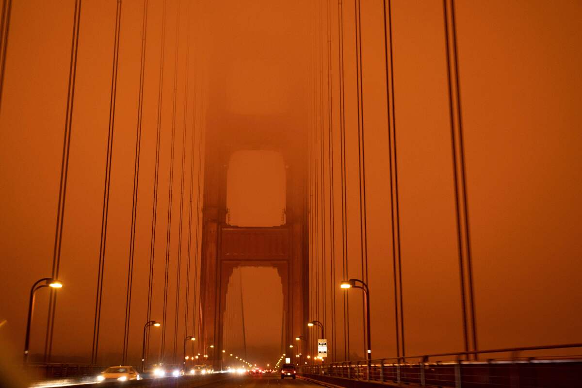 Cars drive across the Golden Gate Bridge with headlights on under a dark orange, smoke-filled sky
