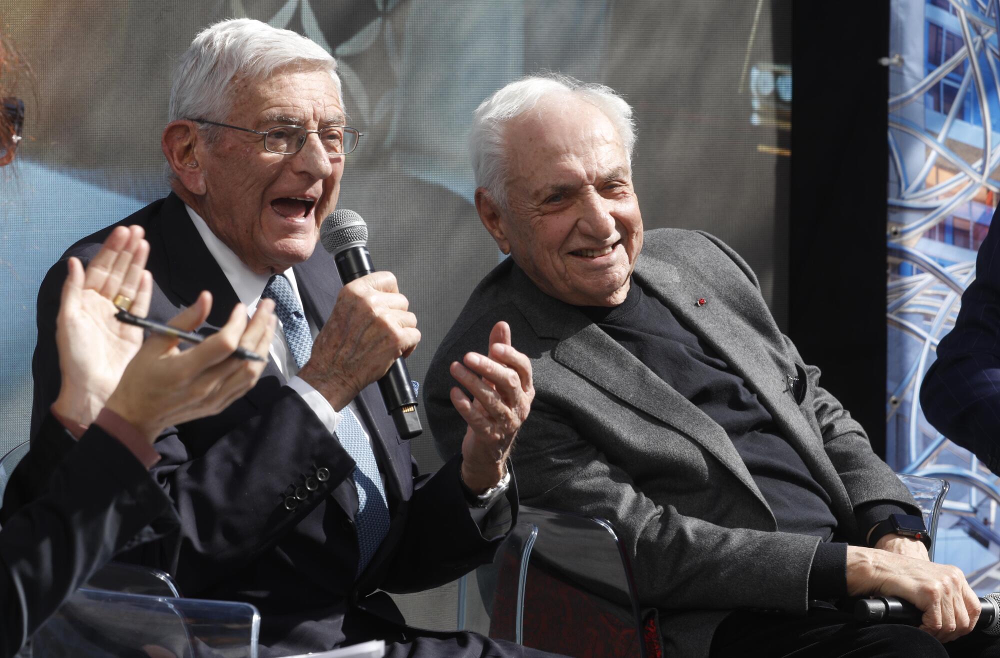 Seated on a stage with Frank Gehry, Eli Broad speaks into a microphone.