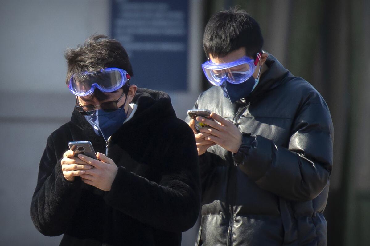 Travelers wear face masks and goggles as they use their smartphones outside the Beijing Railway Station on Saturday in Beijing.