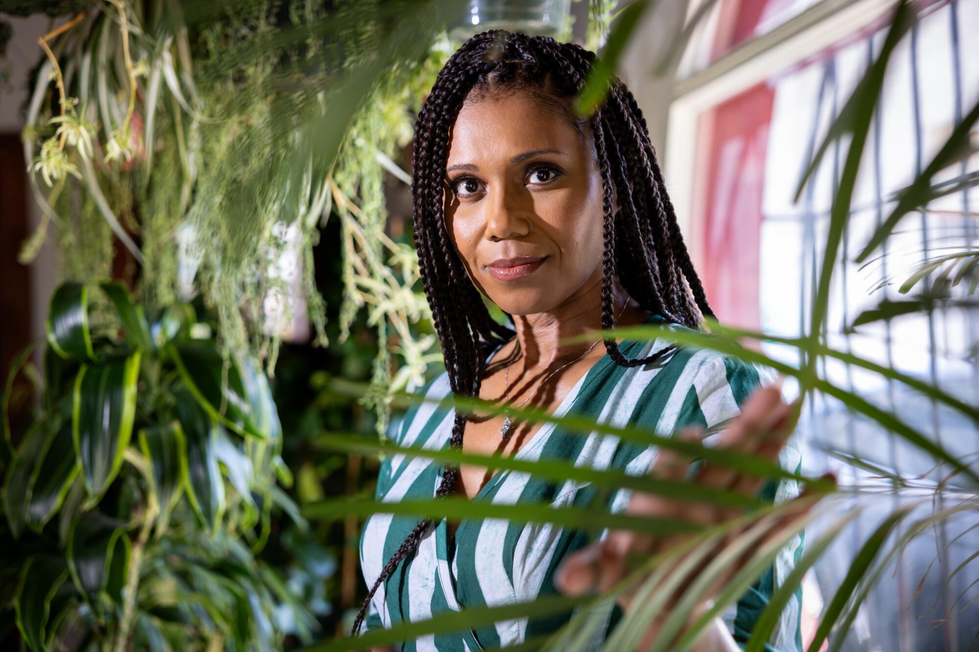 A woman in a blue and green striped dress stands near a window by some plants.