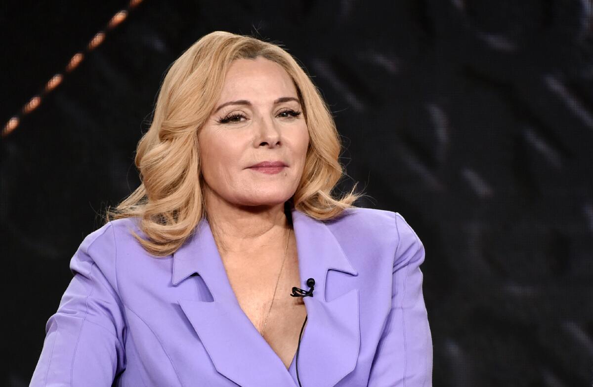 Kim Cattrall seated, wearing a lavender suit 