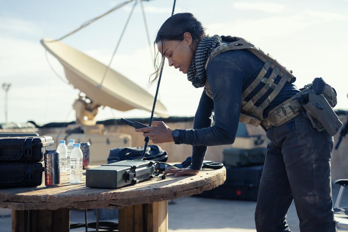 A woman dressed in a protective camo vest leans over a table and looks at a phone. A satellite is in the background.