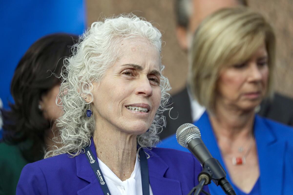 A woman with gray hair, wearing a purple jacket and white top, speaks before a microphone 