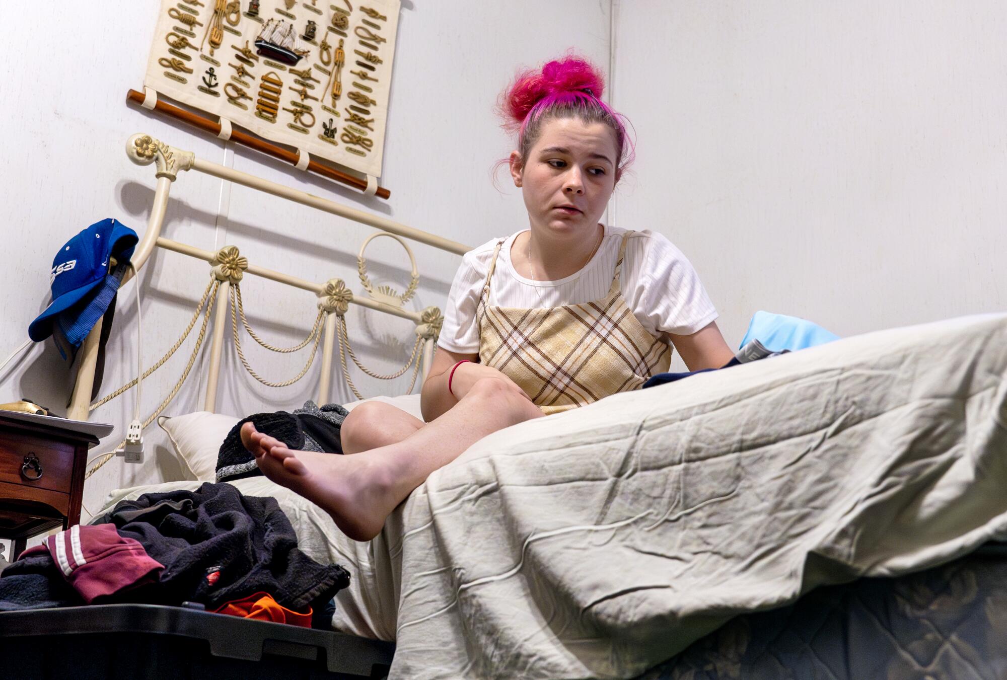Framed  from a low angle, a teen girl sits on a bed, staring off with a forlorn look
