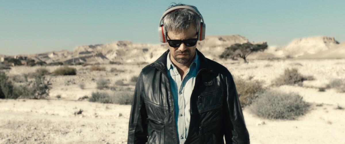 A man in sunglasses and headphones walks through a desert landscape.