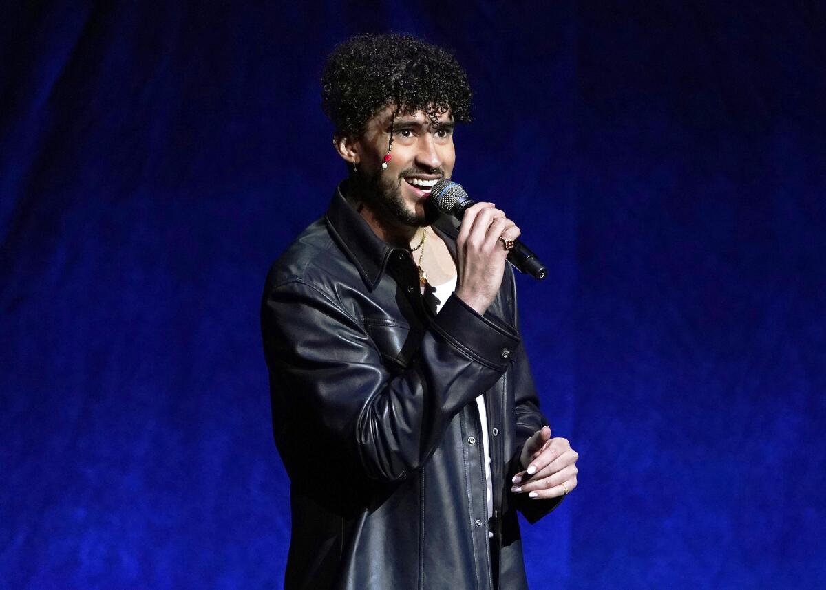 A musical artist in a black leather jacket stands holding a microphone onstage