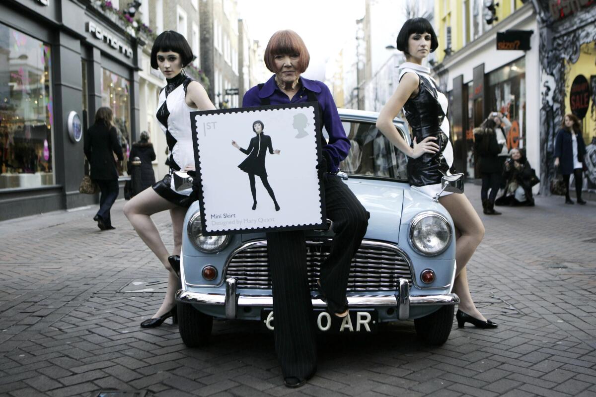 A woman sits on a small car's hood flanked by models and holding a framed photo of a postage stamp featuring another model