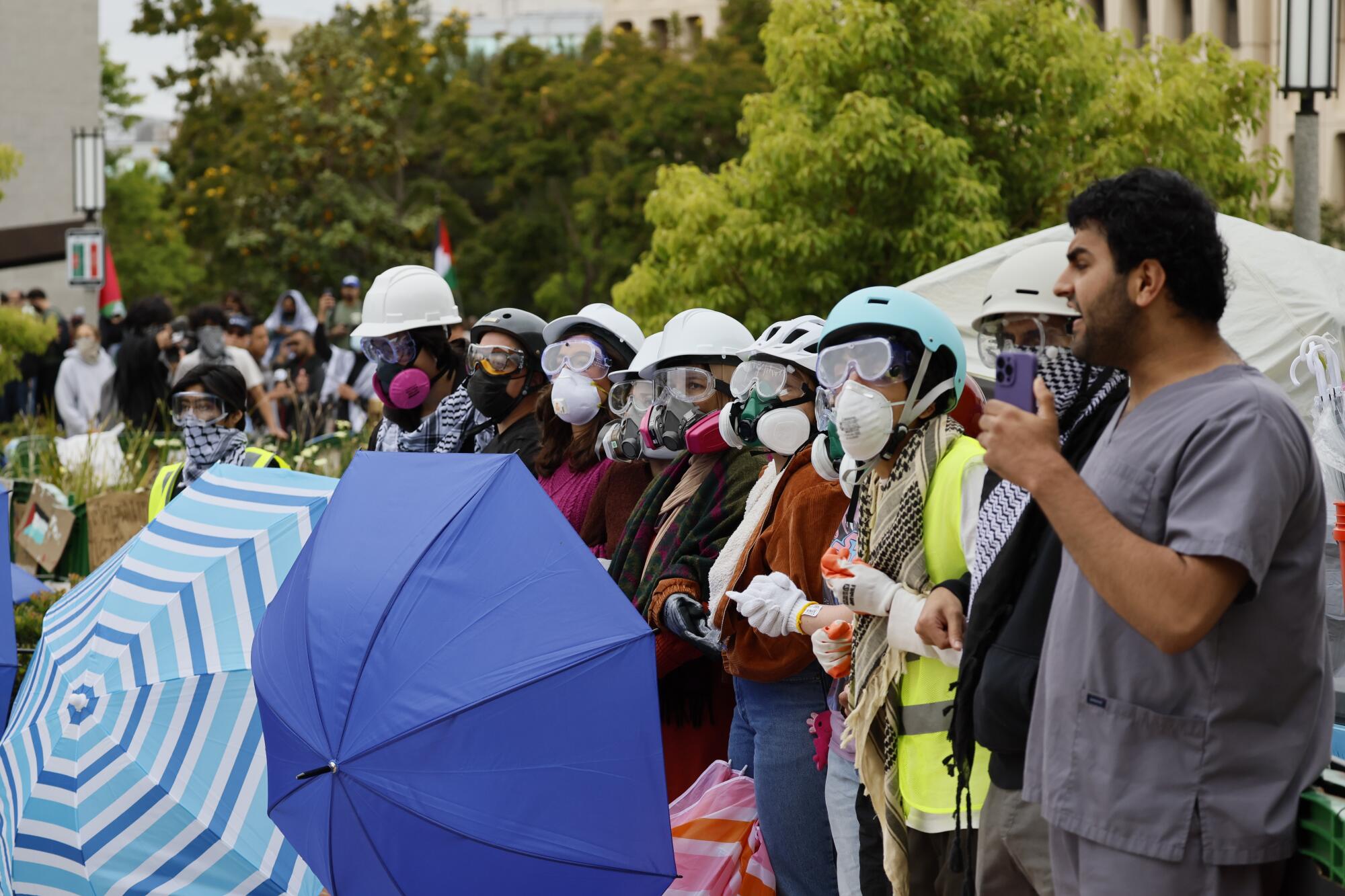 Pro-Palestinian protesters with masks and helmets linking arms to form a blockade, some holding open umbrellas