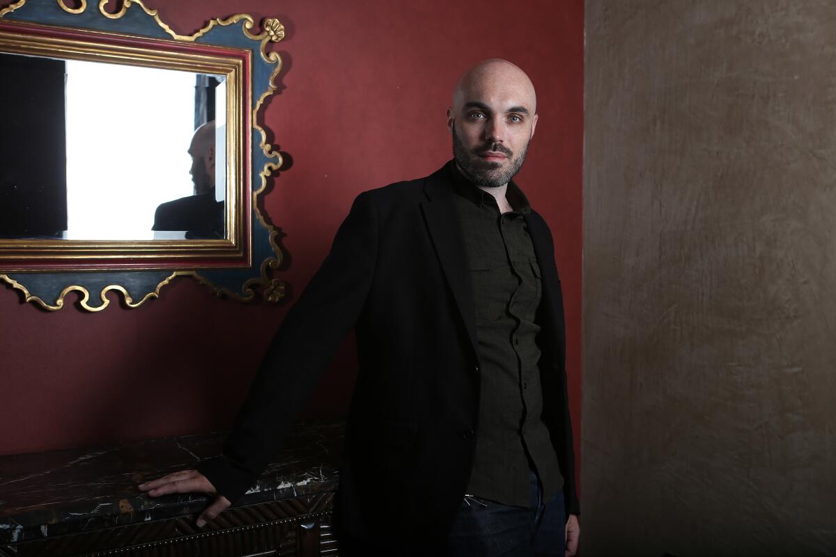 A man with a shaved head and a short beard wears a black shirt and stands before a red wall hung with a framed mirror.