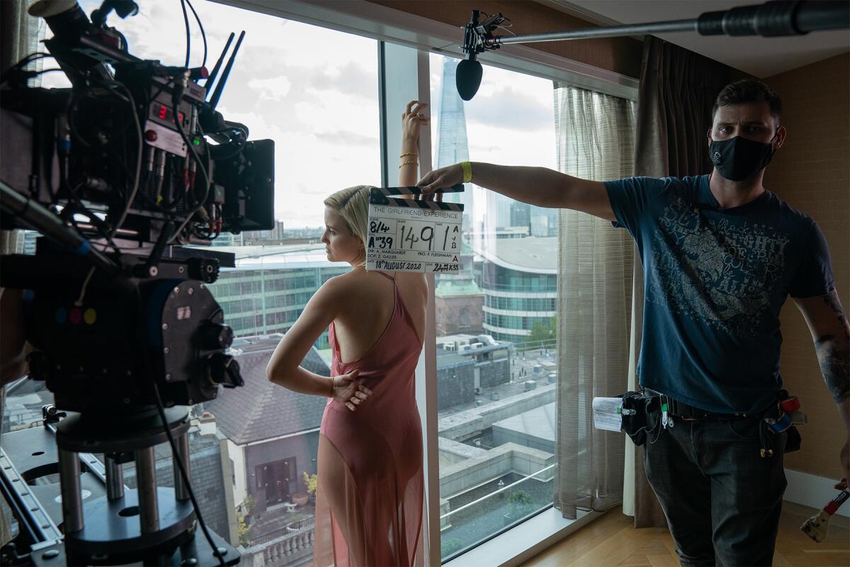 A woman in a sheer pink dress looks out a window, alongside a camera and a man with a film slate.