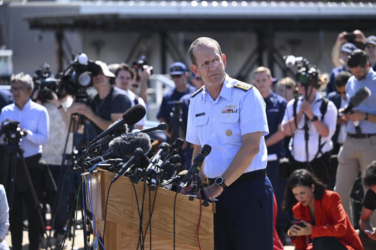A man in a Navy uniform stands outdoors, surrounded by people with film cameras, and looks to right from a lectern with mics
