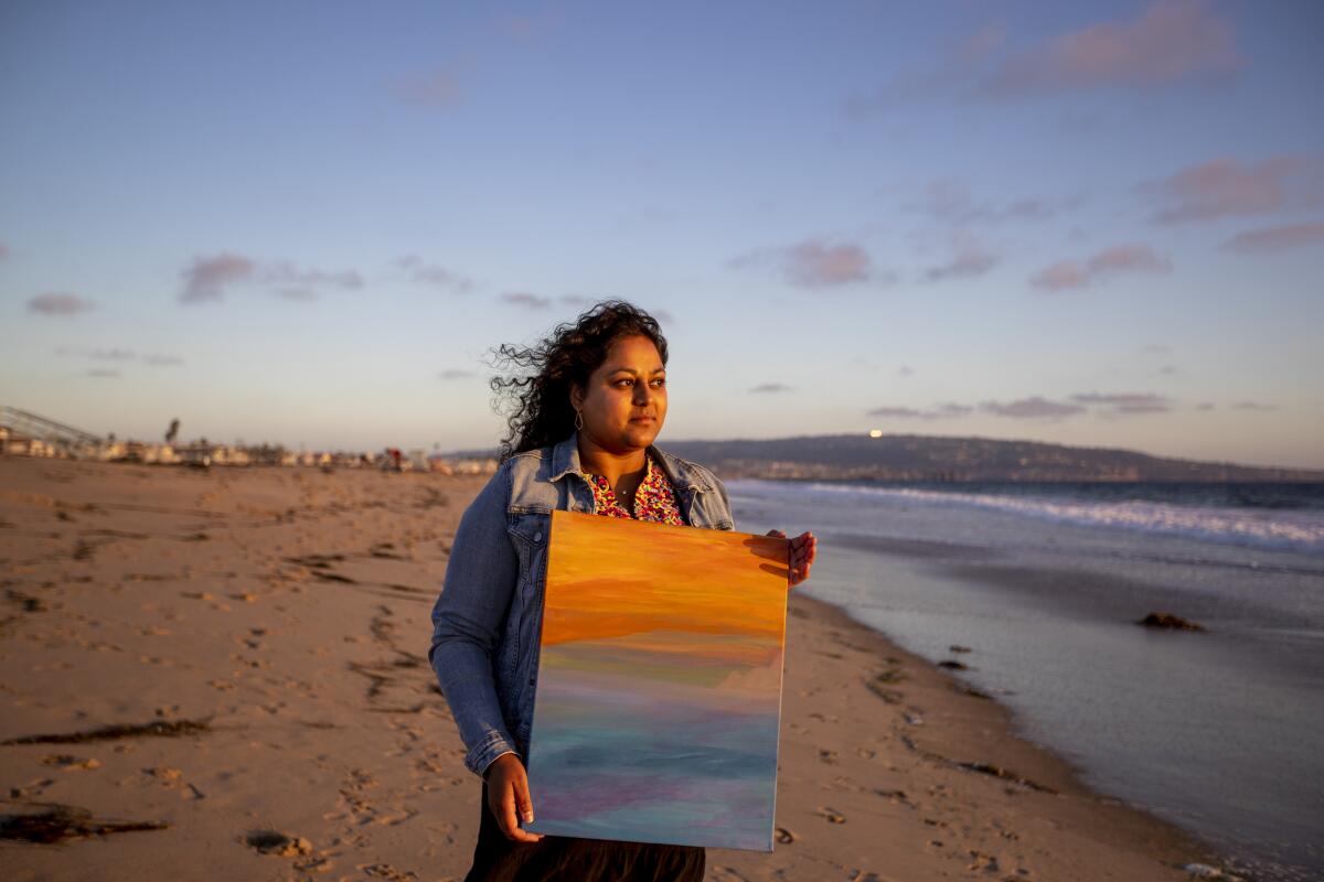 A woman with dark hair standing on a beach holds a painting in orange and blue colors