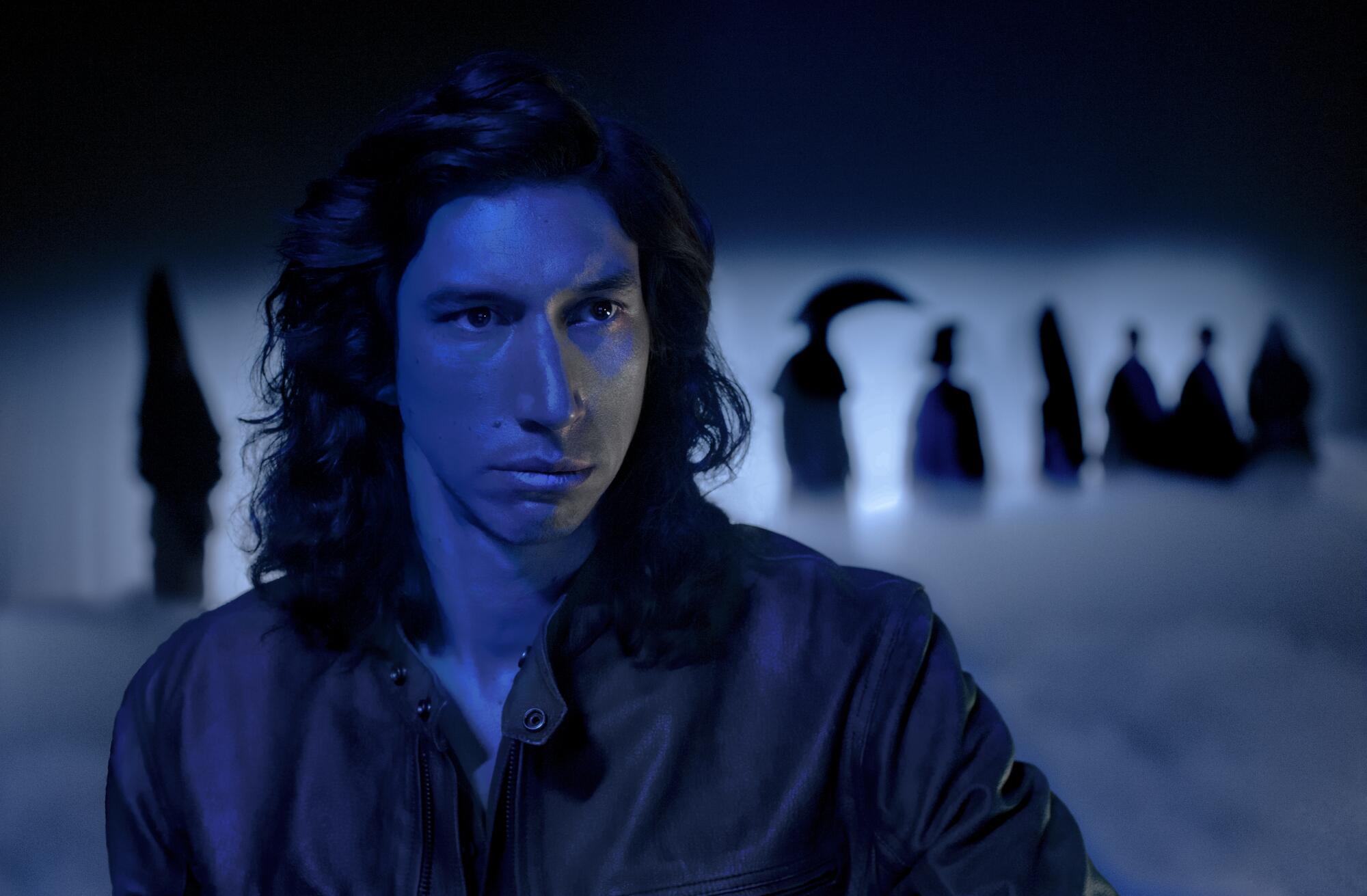Adam Driver, his face lit blue, with shadowy figures behind him, in the movie "Annette."