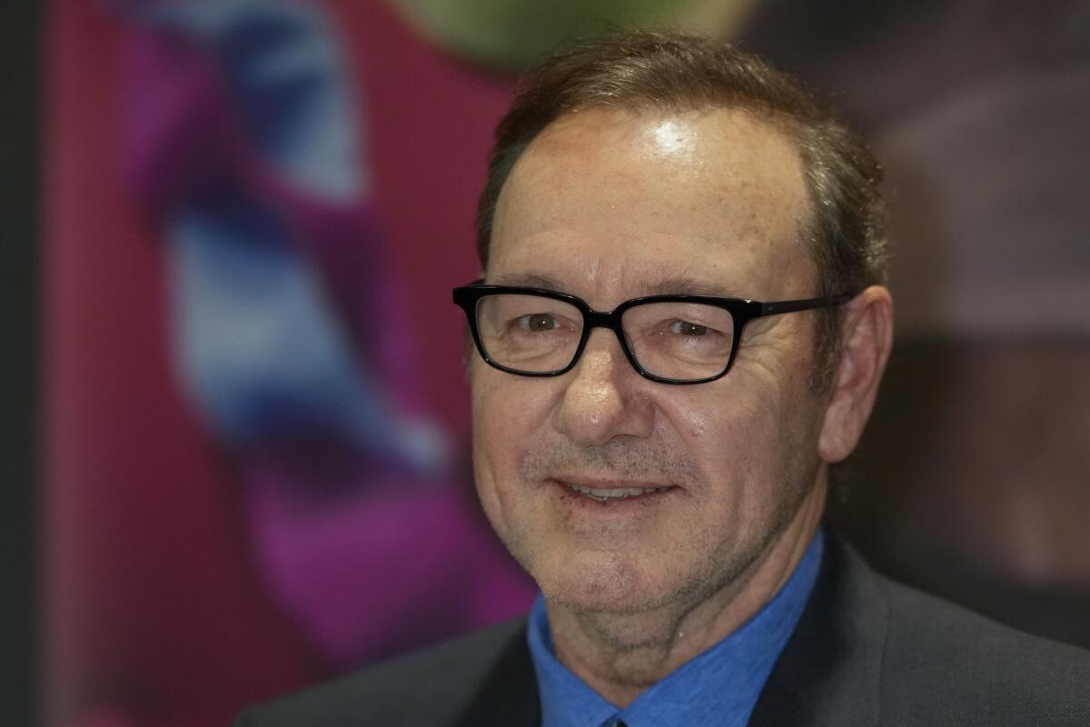 Kevin Spacey smiling and wearing thin-framed black glasses, a blue suit shirt and a dark blazer 