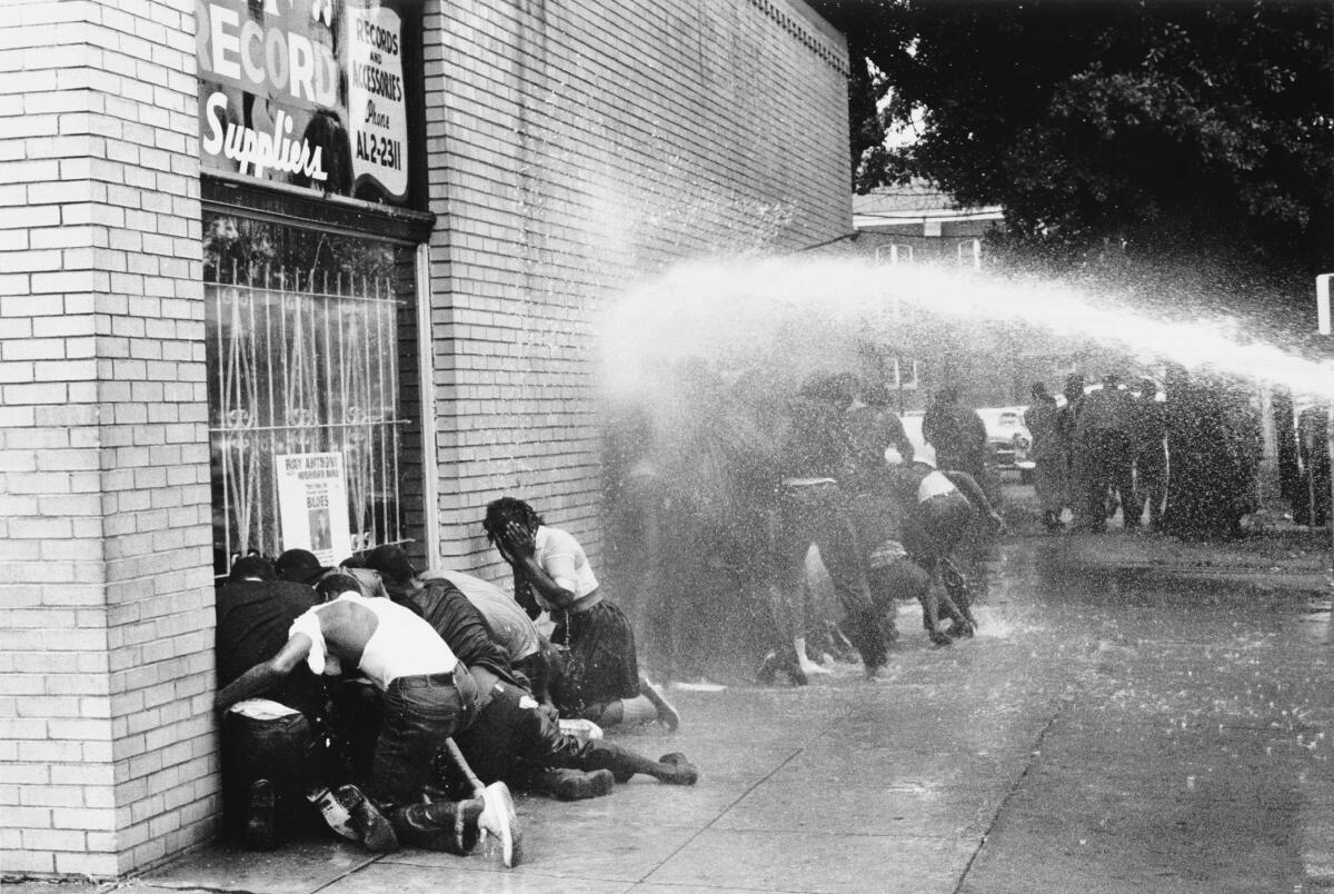 A black-and-white horizontal frame of a street scene — people huddle against a building wall while sprayed by fire hose 