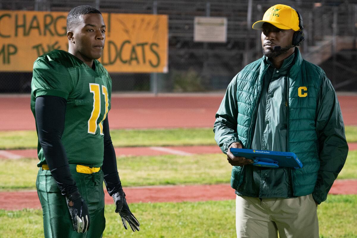 A high school football player stands on the field with his coach, who holds a clipboard.