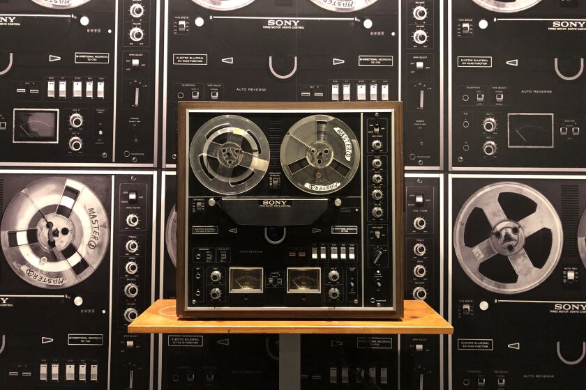 A reel-to-reel tape recorder is seen on a shelf before a wall covered in a wallpaper showing reel-to-reel recorders
