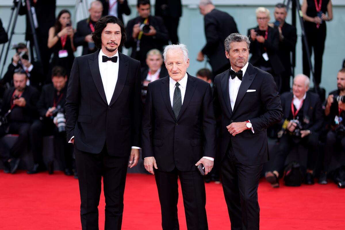 Three men in tuxedos or dark suits stand on a red carpet