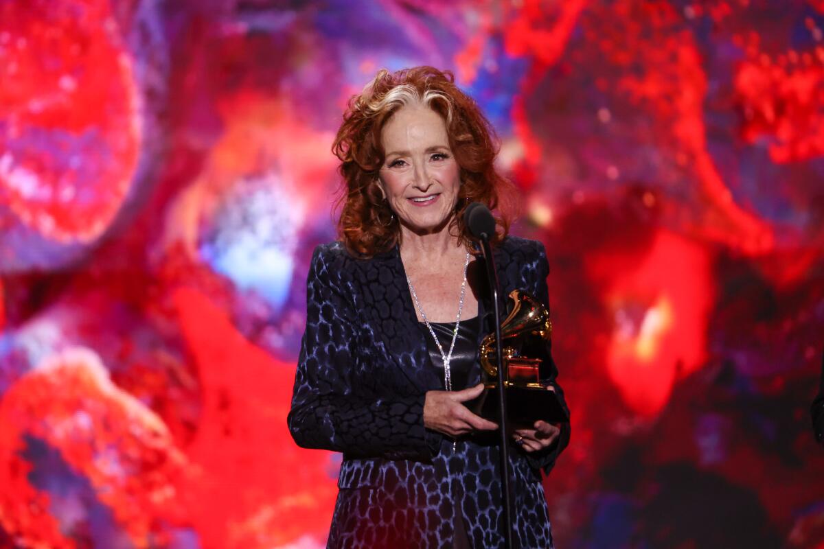 Bonnie Raitt, holding her Grammy, speaks at a microphone.