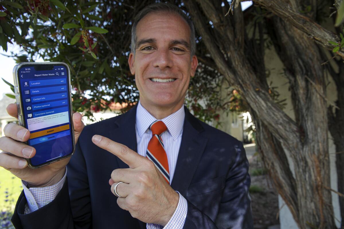 A smiling man in suit and tie holds up a smartphone in one hand and points at the screen with the other.