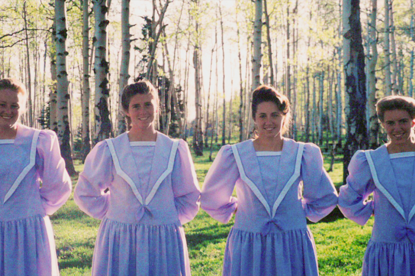 Four women in conservative purple dresses with their hands behind their backs, standing in a wood.
