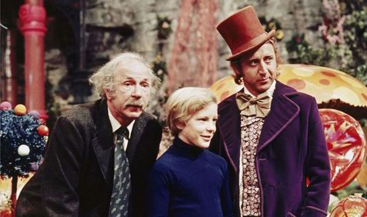 A jaunty man in a top hat and a purple velvet suit, Willy Wonka, stands next to a young boy and his grandfather.