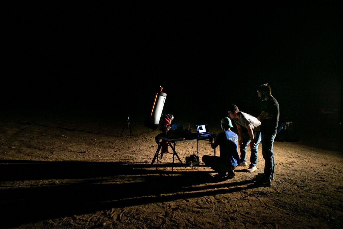 Three people stand near a laptop on a table in the desert, in front of a telescope.