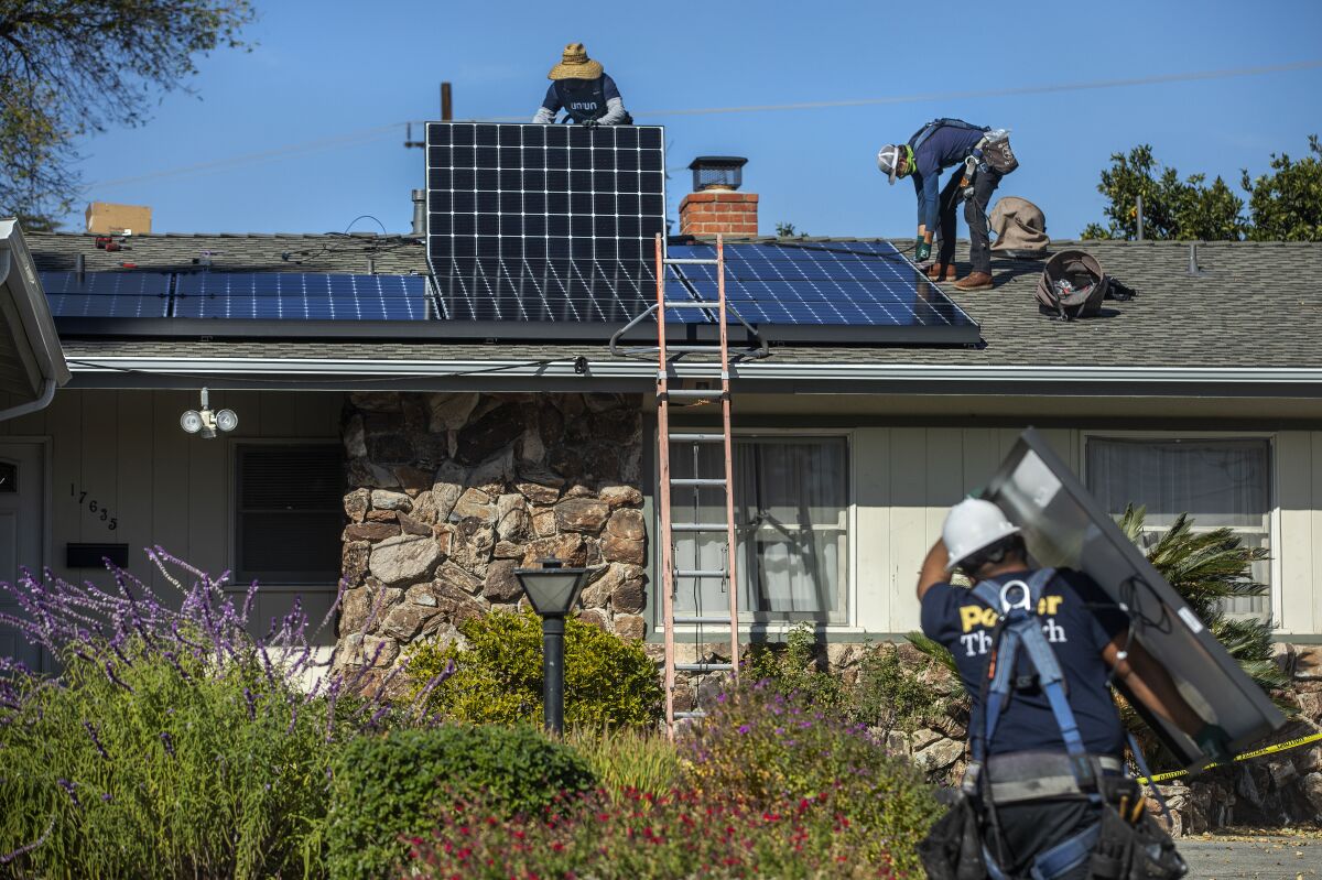 A man carries a solar panel and two others work with tools on a roof to install the panels.