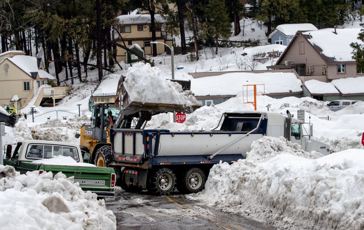 Amid tall banks of snow bordering a roadway, a bulldozer empties a load of snow into a truck.