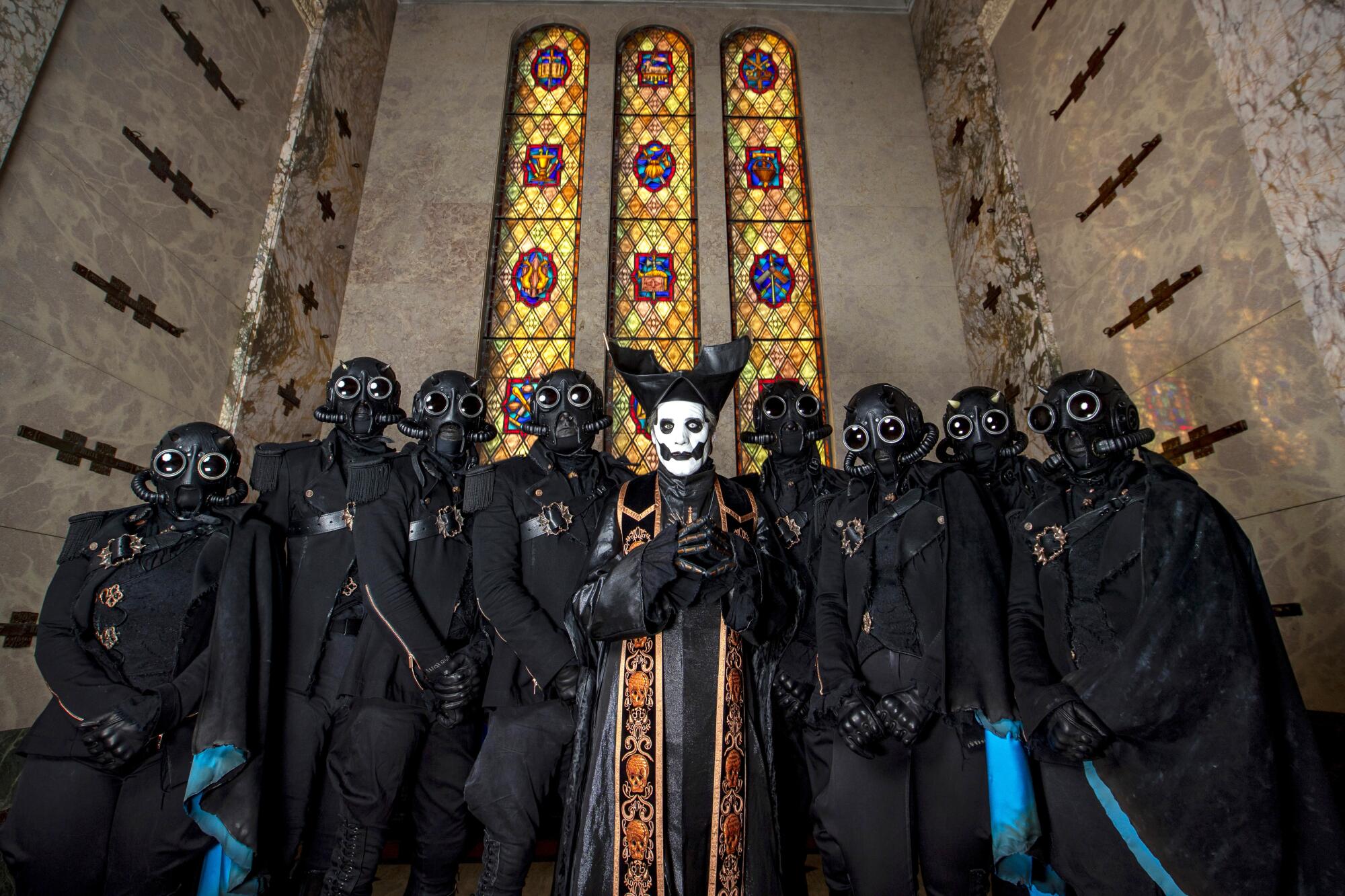 Nine musicians in goth costumes, including eight wearing black gas masks and one wearing a white latex mask