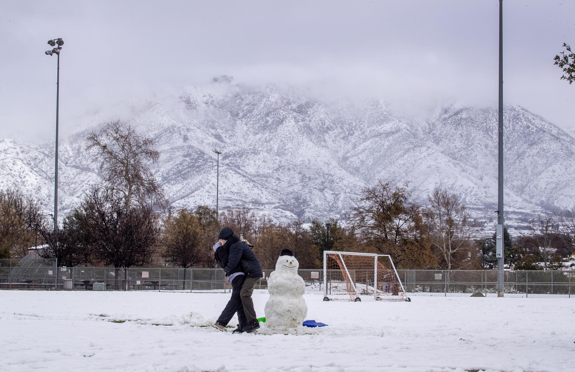 Two people hug next to a snowman on a snow-covered soccer field