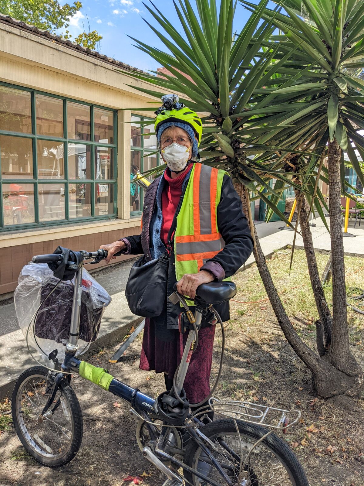 Mary White, wearing a snow cap and reflector vest, stands next to her collapsible bicycle.