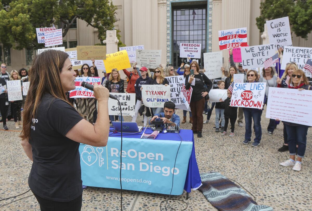 A woman holding a microphone stands in a building plaza before a group of people bearing signs opposing pandemic closures.