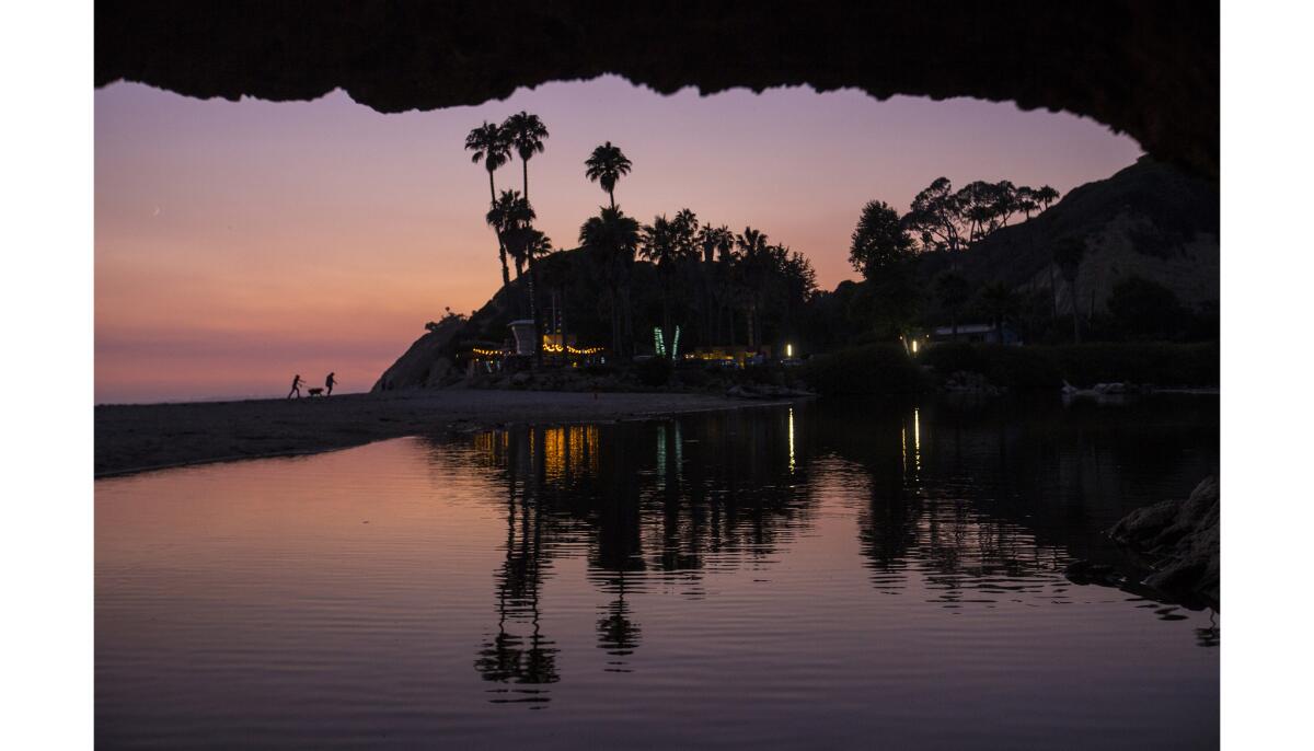 A tidal inlet reflects the surrounding landscape as a couple walk with their dog at twilight along Arroyo Burro Beach County Park in Santa Barbara.