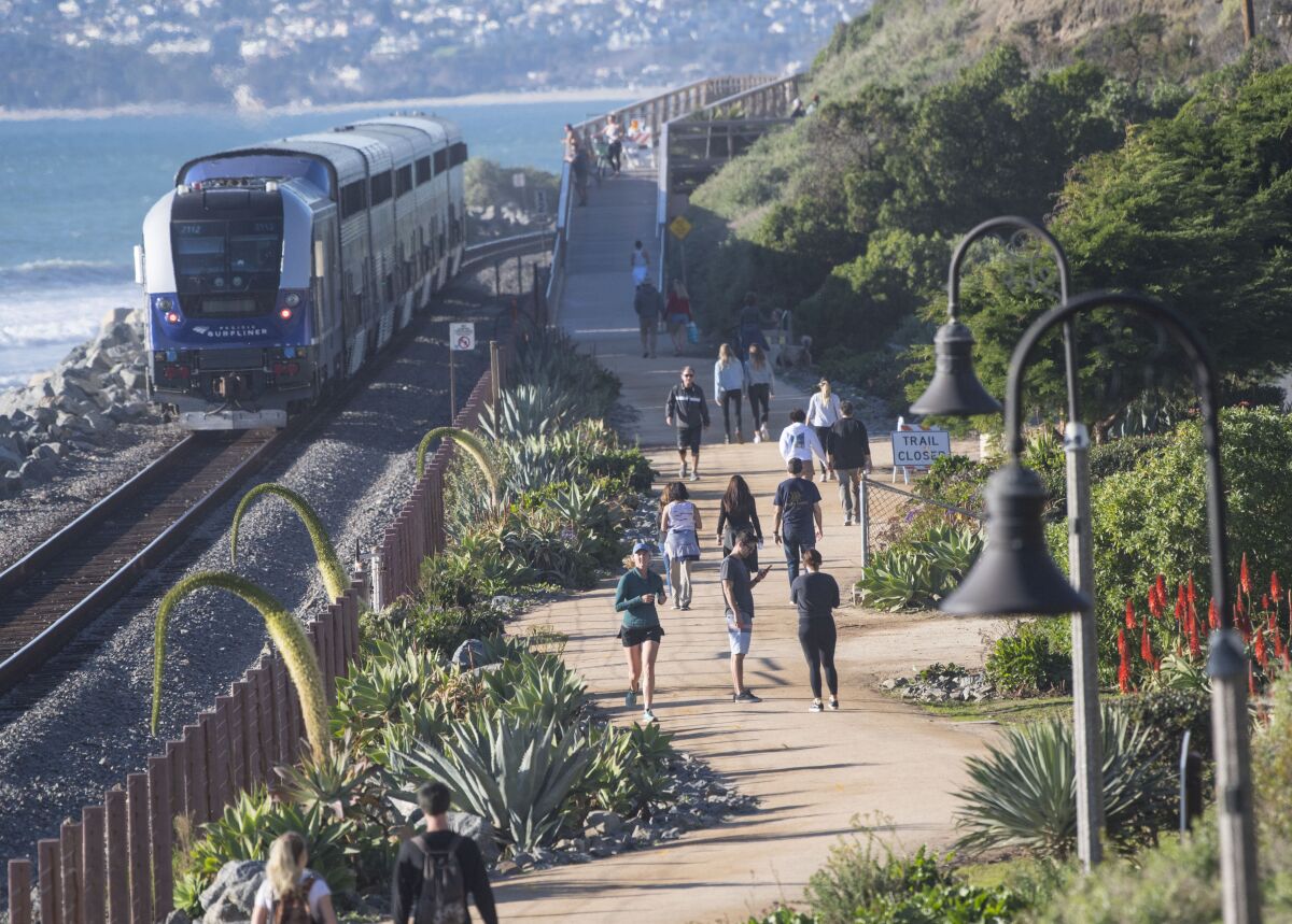 A train hugs the coast while people walk along a trail that runs alongside the tracks