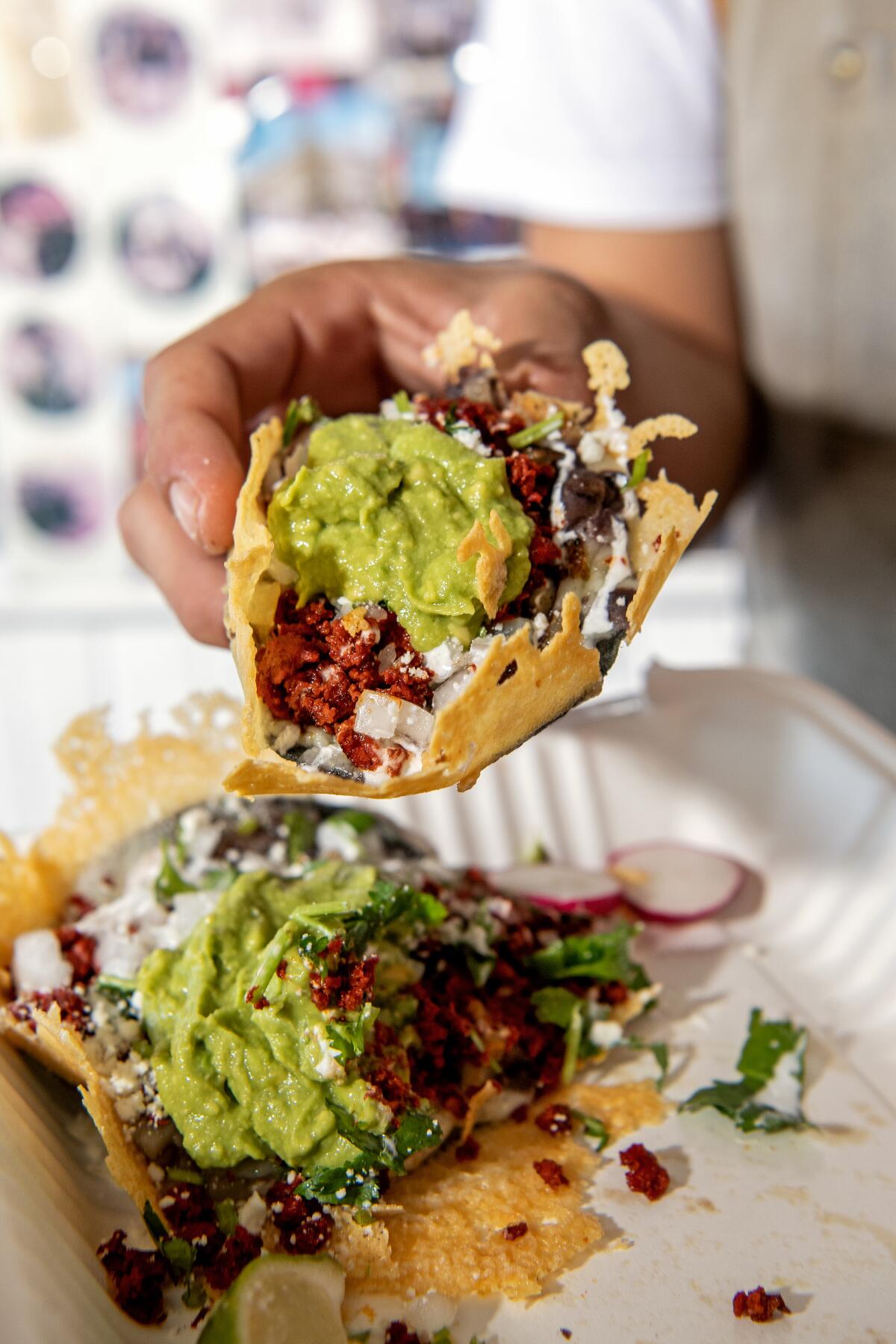 A hand holds a loaded taco with guacamole over a paper plate, on which rests another taco